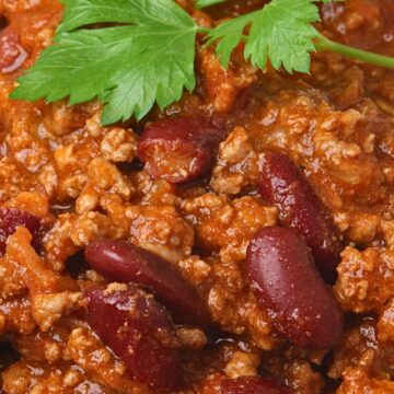 Close-up of high protein beef chili with ground beef, red kidney beans, and thick tomato-based sauce, garnished with fresh green parsley. Hearty, saucy texture with visible beans and beef, served over white rice, highlighting a comforting weeknight chili recipe.