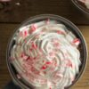 Overhead view of a mug of mint hot chocolate topped with a thick swirl of whipped cream and crushed peppermint candy. Red and white candy cane pieces sprinkled on top. Set on a rustic wooden table with candy canes and another mug partially visible in the background.