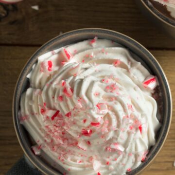 Overhead view of a mug of mint hot chocolate topped with a thick swirl of whipped cream and crushed peppermint candy. Red and white candy cane pieces sprinkled on top. Set on a rustic wooden table with candy canes and another mug partially visible in the background.