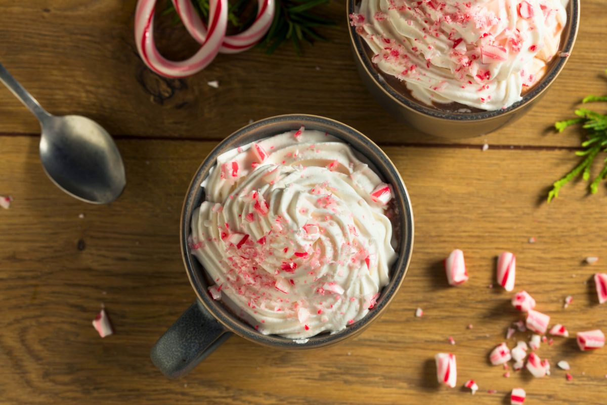 Overhead view of two mugs of mint hot chocolate topped with whipped cream and crushed peppermint candy. Red and white candy cane pieces scattered on top and on a rustic wooden table. A metal spoon, whole candy canes, and evergreen sprigs add a cozy holiday hot chocolate scene.