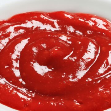 Close-up of thick homemade tequila cocktail sauce in a small white bowl, bright red with a glossy finish and visible swirl marks. Smooth, rich texture with tiny flecks throughout, photographed against a clean white background to highlight color and consistency.