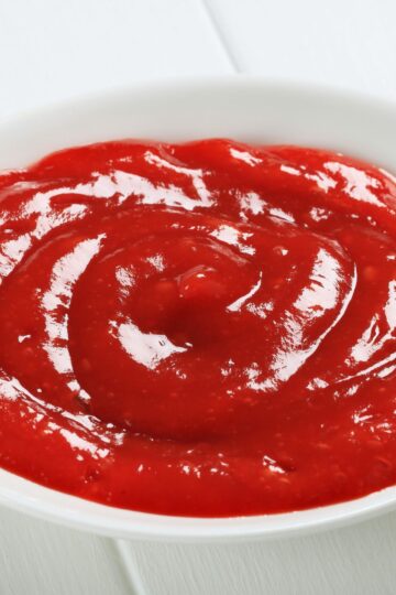 Close-up of thick homemade tequila cocktail sauce in a small white bowl, bright red with a glossy finish and visible swirl marks. Smooth, rich texture with tiny flecks throughout, photographed against a clean white background to highlight color and consistency.