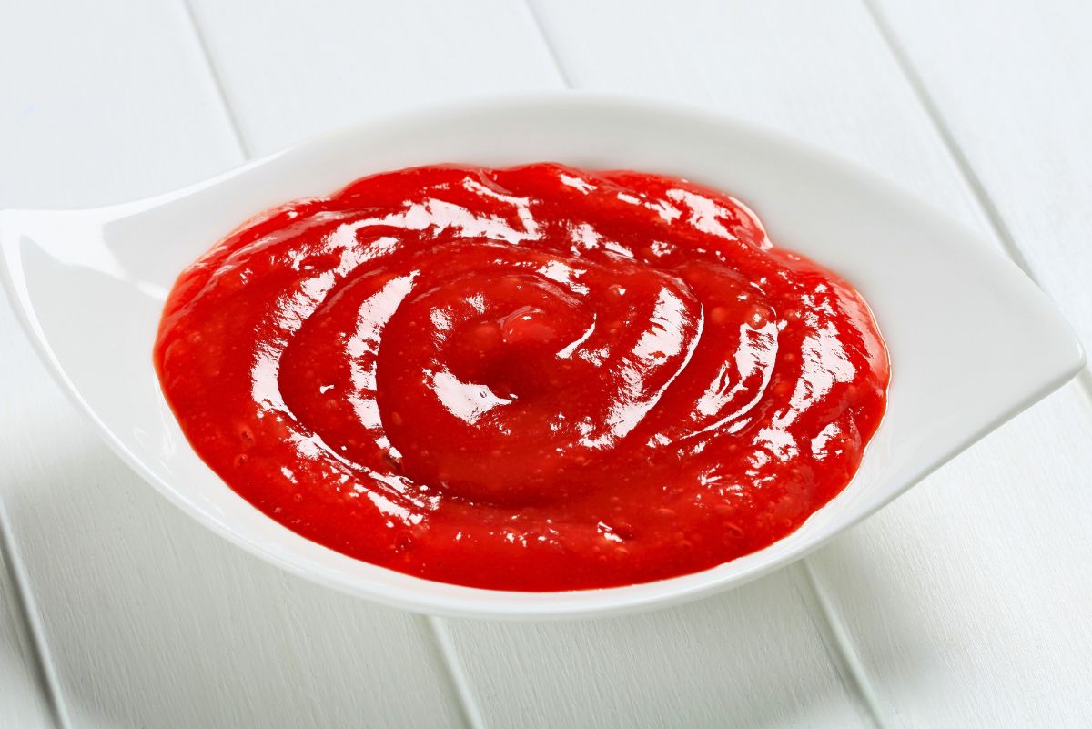 Bright red homemade tequila cocktail sauce swirled smoothly in a small white serving dish. Glossy, thick texture with visible ridges from stirring, set on a white wooden surface in clean, minimal food photography.