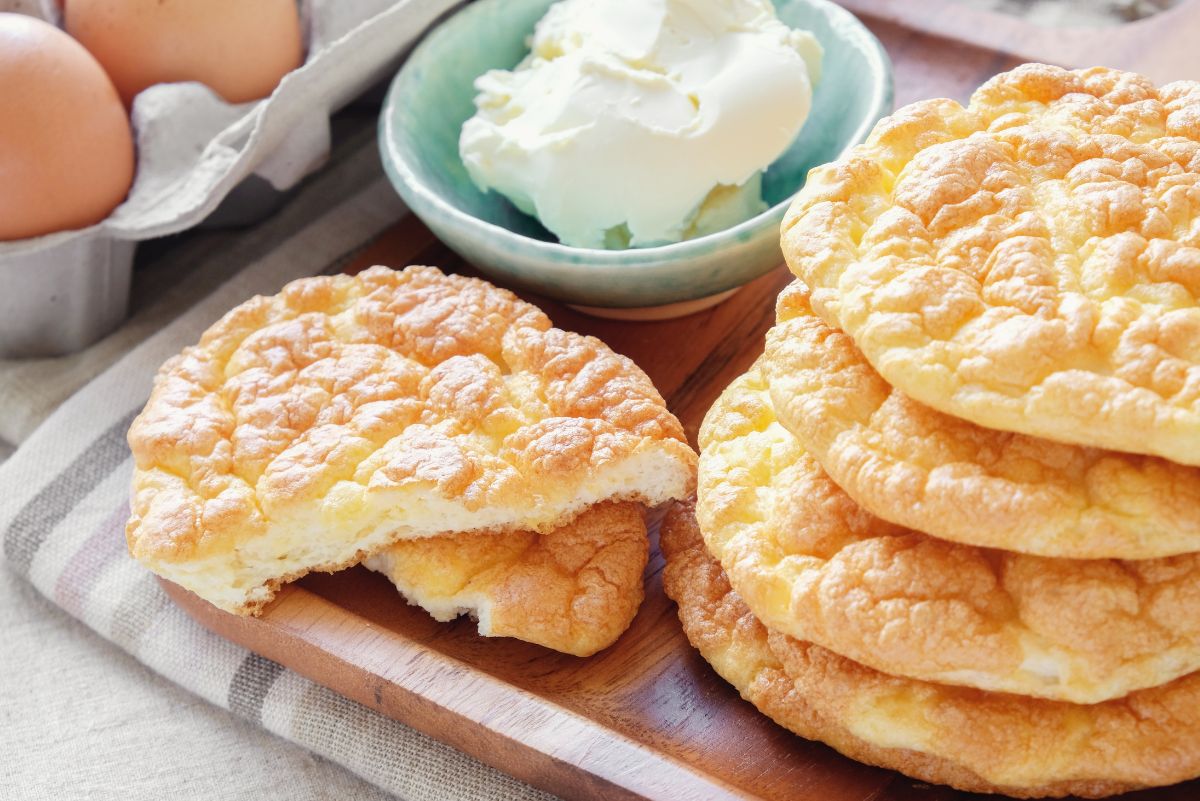 Low carb cloud bread rounds stacked on a wooden board, golden and fluffy with a light, airy texture. One piece is broken open to show the soft interior. Eggs in a carton and a small bowl of cream cheese sit nearby, highlighting simple ingredients used for cloud bread.