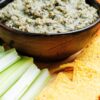 Creamy black bean dip in a dark bowl on a wooden board, thick and slightly chunky with visible herbs and beans. Served with golden tortilla chips and fresh celery sticks, styled as a quick healthy appetizer in close-up food photography.