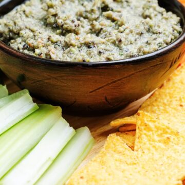 Creamy black bean dip in a dark bowl on a wooden board, thick and slightly chunky with visible herbs and beans. Served with golden tortilla chips and fresh celery sticks, styled as a quick healthy appetizer in close-up food photography.