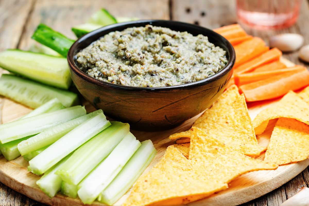Creamy black bean dip in a dark bowl on a wooden board, thick and slightly chunky with visible herbs. Served with celery sticks, carrot sticks, and crispy tortilla chips, styled as a healthy quick appetizer with fresh vegetables and crunchy chips.