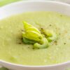 Creamy celery soup in a white bowl, pale green and smooth with a velvety texture. Garnished with sliced celery, a small celery leaf, and cracked black pepper on top. Fresh celery stalks blurred in the background on a light kitchen surface, highlighting homemade cream of celery soup.
