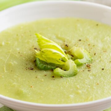 Creamy celery soup in a white bowl, pale green and smooth with a velvety texture. Garnished with sliced celery, a small celery leaf, and cracked black pepper on top. Fresh celery stalks blurred in the background on a light kitchen surface, highlighting homemade cream of celery soup.
