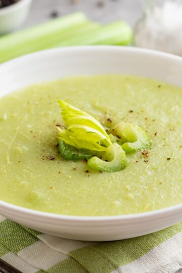 Creamy celery soup in a white bowl, pale green and smooth with a velvety texture. Garnished with sliced celery, a small celery leaf, and cracked black pepper on top. Fresh celery stalks blurred in the background on a light kitchen surface, highlighting homemade cream of celery soup.