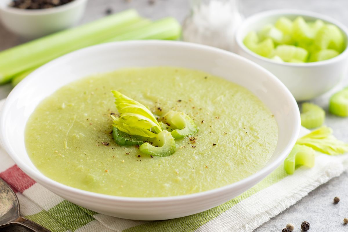 Creamy celery soup in a white bowl, smooth and pale green with a velvety texture. Garnished with sliced celery, a small celery leaf, and cracked black pepper. Fresh celery stalks and chopped celery pieces sit nearby on a light kitchen surface, showing homemade cream of celery soup.
