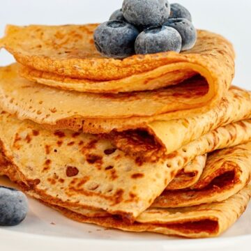 Close-up of folded savory chickpea flour crepes stacked on a white plate, lightly browned with crisp edges and soft layers. Fresh blueberries sit on top and nearby, adding contrast. Clean white background with bright, minimal food photography highlighting texture and color.
