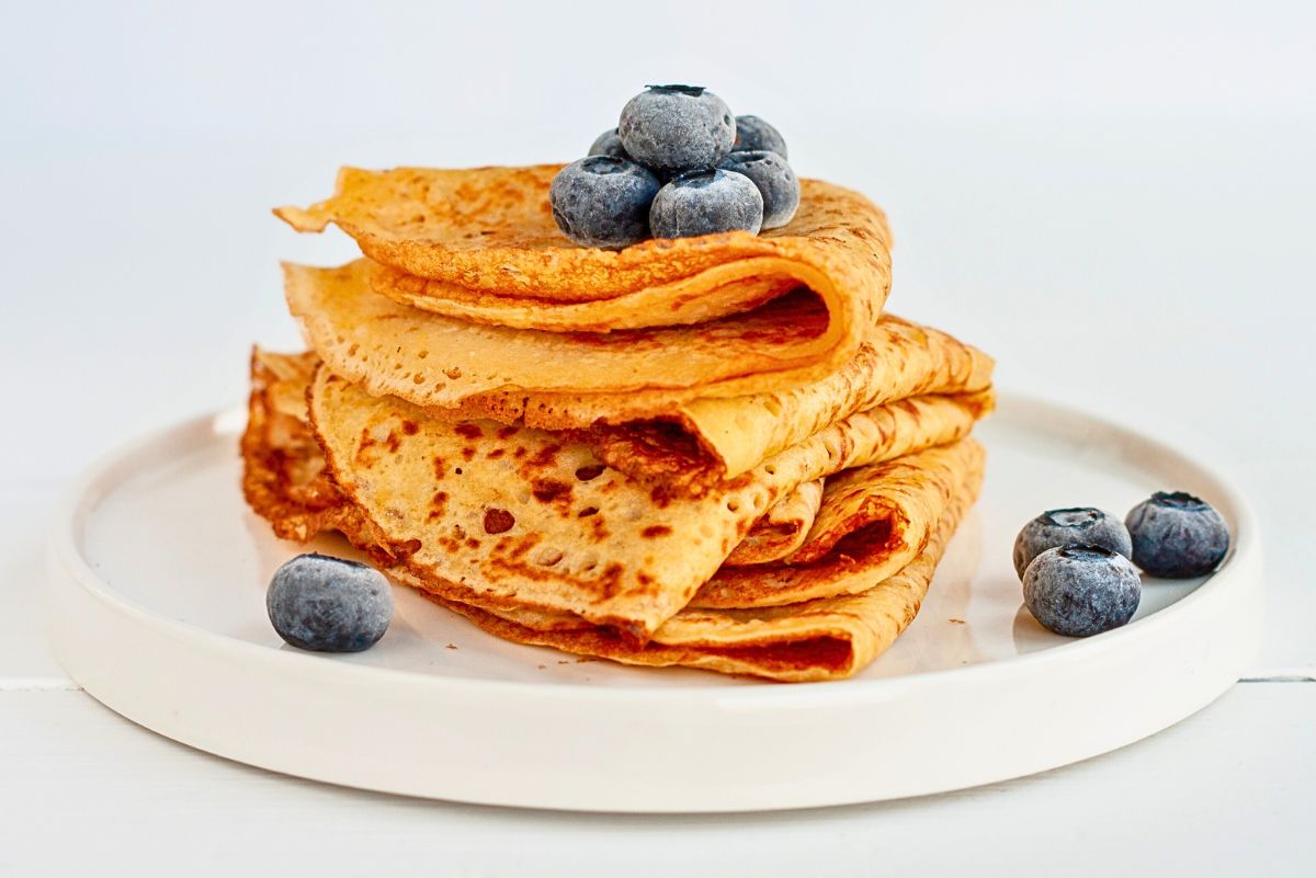 Stack of golden savory chickpea flour crepes folded neatly on a white plate, lightly browned with crisp edges and soft layers. Fresh blueberries sit on top and around the plate. Clean white background with bright food photography highlighting texture and color.