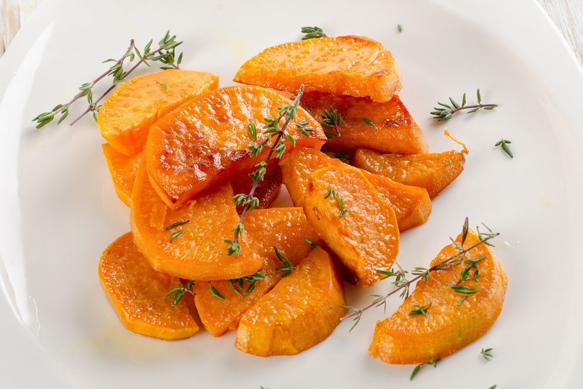 Thyme roasted sweet potato wedges arranged on a white plate, lightly crisped and golden with a glossy roasted finish. Fresh thyme sprigs scattered over the tender orange wedges, showing caramelized edges and a soft interior in bright, clean food photography.