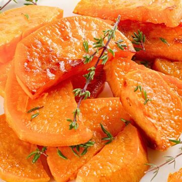 Thyme roasted sweet potato wedges piled on a white plate, golden and glossy with lightly crisped edges. Fresh thyme sprigs scattered over tender orange wedges, showing a soft interior and roasted surface in close-up food photography.