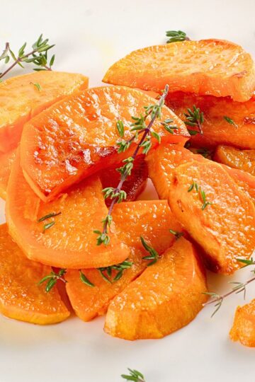 Thyme roasted sweet potato wedges piled on a white plate, golden and glossy with lightly crisped edges. Fresh thyme sprigs scattered over tender orange wedges, showing a soft interior and roasted surface in close-up food photography.