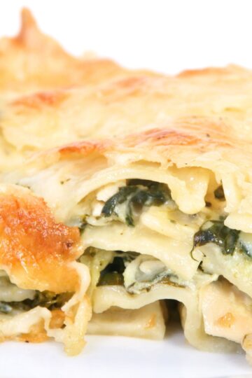 Close-up of white sauce chicken spinach lasagna with layers of tender lasagna noodles, creamy white sauce, shredded chicken, and wilted spinach. The top is covered in melted golden cheese with lightly browned edges, showing a rich, cheesy texture and creamy filling inside.