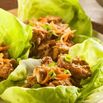 Close-up of Asian chicken lettuce wraps with crisp green lettuce leaves filled with tender chicken pieces, shredded carrots, and sliced green onions. Fresh lettuce cups arranged on a white plate with a warm, casual presentation.