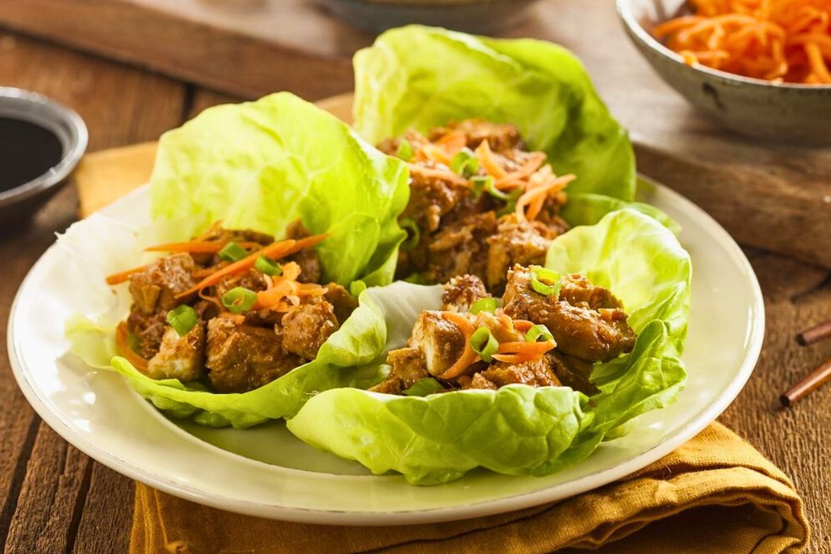 Asian chicken lettuce wraps served on a white plate, with crisp green lettuce cups filled with tender chicken, shredded carrots, and sliced green onions. Fresh, colorful lettuce wraps styled on a rustic wooden table with dipping sauce and carrots nearby.
