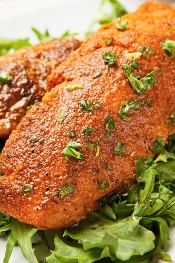 Blackened salmon fillet with a deep golden brown spice crust resting on mixed leafy greens. The salmon is coated in seasoning, lightly glossy with oil, and sprinkled with chopped fresh herbs, served as a high protein salmon salad on a white plate.