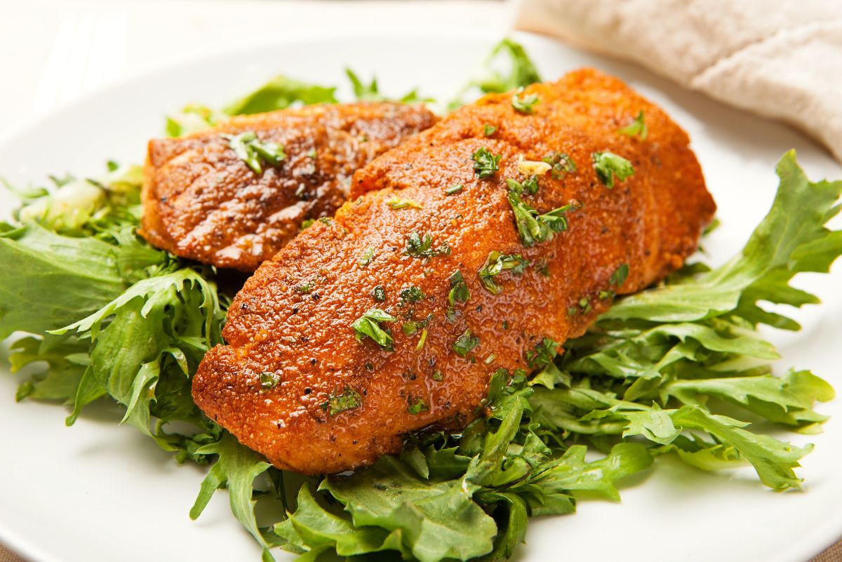 Blackened salmon fillets with a deep spiced crust served over mixed leafy greens on a white plate. The salmon is glossy with oil and sprinkled with chopped fresh herbs, resting on fresh green lettuce as a high protein salmon salad.