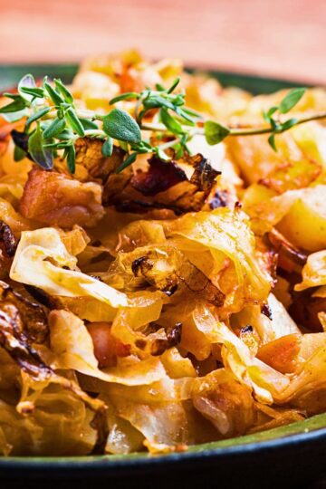 Close-up of a cabbage skillet with bacon and potatoes, showing golden caramelized cabbage, crispy bacon pieces, and tender potato chunks cooked in a pan, topped with fresh green herb sprigs. Rustic skillet dinner with savory cabbage and potatoes.