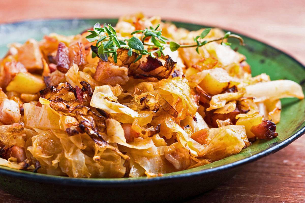 Cabbage skillet with bacon and potatoes served in a green bowl, showing caramelized cabbage, crispy bacon pieces, and tender potato chunks cooked until golden, topped with fresh thyme. Rustic comfort food skillet meal with cabbage and potatoes.