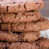 Close-up stack of thick chocolate almond cookies with a soft, slightly cracked surface and visible sugar crystals. The cookies are rich brown, chewy-looking, and layered on a rustic wooden surface, highlighting their dense texture and chocolate cayenne spice baked finish.
