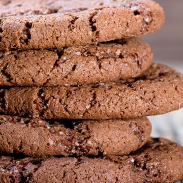 Close-up stack of thick chocolate almond cookies with a soft, slightly cracked surface and visible sugar crystals. The cookies are rich brown, chewy-looking, and layered on a rustic wooden surface, highlighting their dense texture and chocolate cayenne spice baked finish.