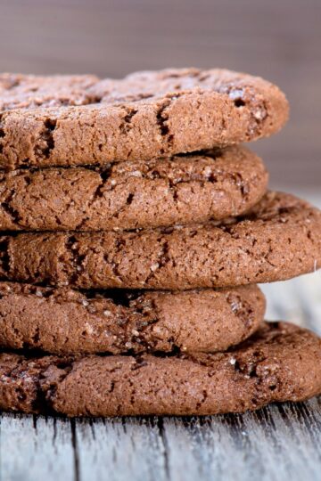 Close-up stack of thick chocolate almond cookies with a soft, slightly cracked surface and visible sugar crystals. The cookies are rich brown, chewy-looking, and layered on a rustic wooden surface, highlighting their dense texture and chocolate cayenne spice baked finish.