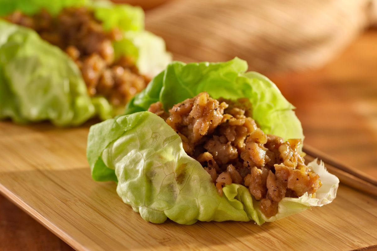 Ginger garlic chicken lettuce wraps arranged on a wooden cutting board, filled with tender chopped chicken coated in a savory ginger garlic sauce. Crisp green butter lettuce leaves cradle the juicy chicken, creating a fresh, healthy, high protein chicken lettuce wrap.