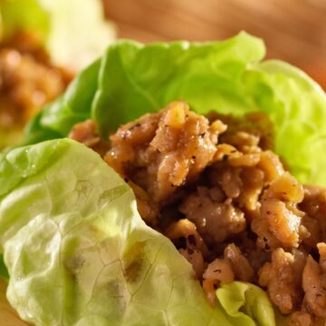 Close-up of ginger garlic chicken lettuce wraps with tender chopped chicken coated in a glossy ginger garlic sauce, nestled in crisp green butter lettuce leaves on a wooden cutting board. Fresh, healthy, high protein chicken lettuce wraps with juicy texture and vibrant green lettuce.