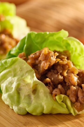 Close-up of ginger garlic chicken lettuce wraps with tender chopped chicken coated in a glossy ginger garlic sauce, nestled in crisp green butter lettuce leaves on a wooden cutting board. Fresh, healthy, high protein chicken lettuce wraps with juicy texture and vibrant green lettuce.