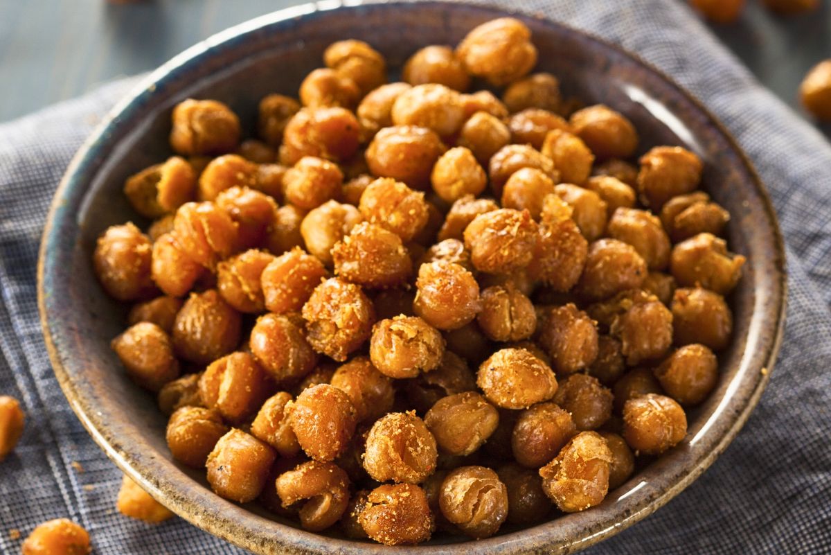 Bowl of crispy roasted chickpeas coated in seasoning, golden brown and crunchy, served on a folded gray cloth on a rustic table. Close-up food photography highlights the textured spice coating and toasted chick peas, with a few scattered in the background. Healthy roasted seasoned chickpeas snack.