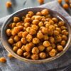 Bowl of crispy roasted seasoned chickpeas, golden brown with a crunchy texture, lightly coated in spices. The chickpeas sit in a rustic ceramic bowl on a gray cloth, with close-up detail showing toasted edges and seasoning. Healthy roasted chickpeas snack.
