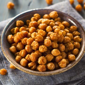 Bowl of crispy roasted seasoned chickpeas, golden brown with a crunchy texture, lightly coated in spices. The chickpeas sit in a rustic ceramic bowl on a gray cloth, with close-up detail showing toasted edges and seasoning. Healthy roasted chickpeas snack.