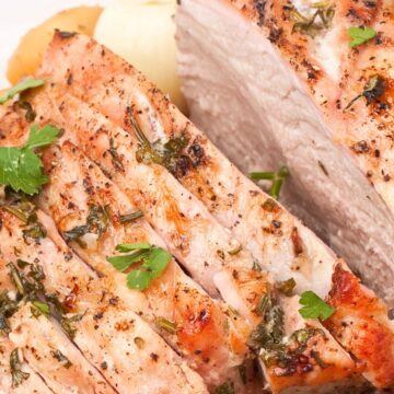 Close-up of herb pepper pork loin roast sliced into thick pieces, showing a juicy interior with a golden browned crust. Garlic, cracked black pepper, and chopped fresh herbs are visible on the pork, served on a white platter with roasted potato slices and parsley garnish.