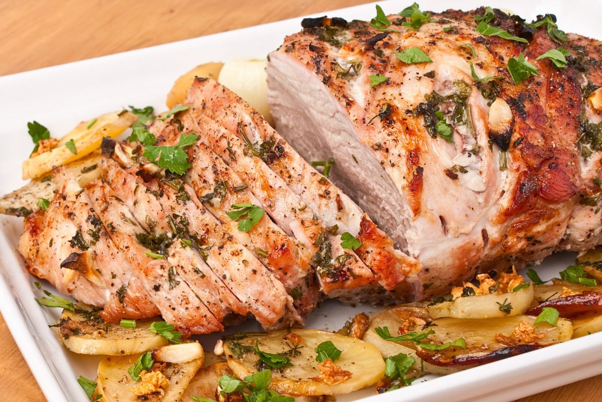Sliced herb pepper pork loin roast with a golden brown crust, visible garlic, cracked black pepper, and chopped fresh herbs. Juicy pork slices arranged on a white serving platter with roasted potato slices and parsley garnish on a wooden surface.