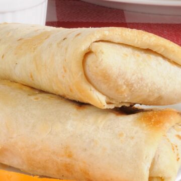 High protein turkey wraps folded and lightly toasted, stacked on a white plate with melted cheese visible at the ends. A small white ramekin of dipping sauce sits in the background on a red checkered cloth.