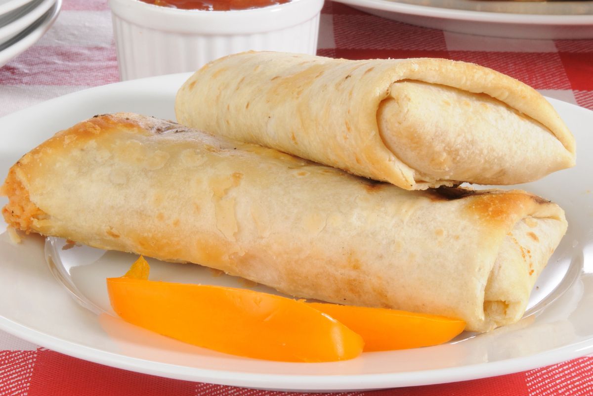 High protein turkey wraps lightly toasted and stacked on a white plate, with melted cheese visible at the ends. Orange bell pepper slices rest beside the wraps. A small white ramekin of dipping sauce sits in the background on a red checkered tablecloth.