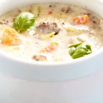 Close-up of high protein crockpot cheeseburger soup in a white ceramic bowl, with creamy broth, ground beef, diced carrots, potatoes, and celery, topped with fresh basil leaves. Bright, soft background highlights the thick, comforting texture of the cheeseburger soup.