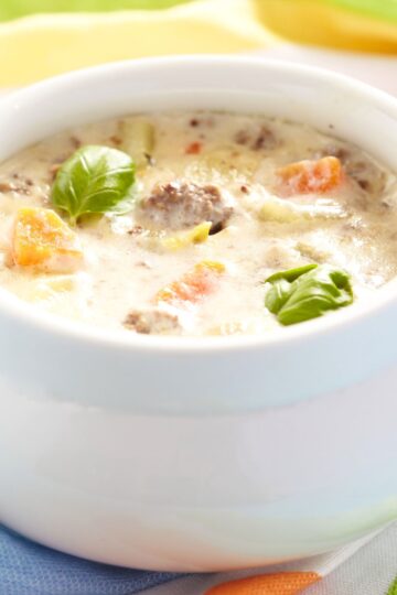 Close-up of high protein crockpot cheeseburger soup in a white ceramic bowl, with creamy broth, ground beef, diced carrots, potatoes, and celery, topped with fresh basil leaves. Bright, soft background highlights the thick, comforting texture of the cheeseburger soup.