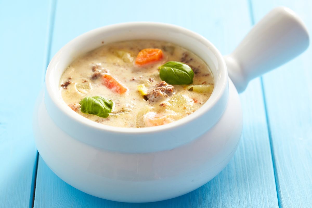 High protein crockpot cheeseburger soup served in a white handled soup bowl on a blue wooden table. Creamy cheeseburger soup with visible ground beef, diced carrots, potatoes, and celery, topped with fresh basil leaves. Bright, clean food photography highlighting a thick, comforting texture.