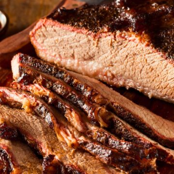 Close-up of sliced Instant Pot BBQ brisket on a wooden cutting board, showing tender beef with a dark caramelized barbecue crust and moist interior. Thick slices reveal smoky bark, juicy texture, and rich glaze, with a knife resting nearby for serving.