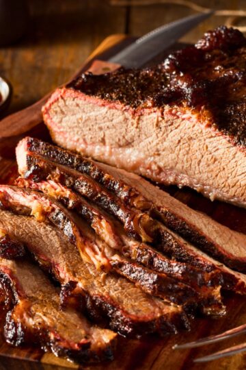 Close-up of sliced Instant Pot BBQ brisket on a wooden cutting board, showing tender beef with a dark caramelized barbecue crust and moist interior. Thick slices reveal smoky bark, juicy texture, and rich glaze, with a knife resting nearby for serving.