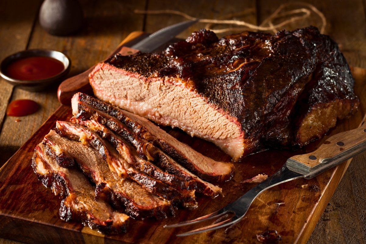 High protein Instant Pot BBQ brisket sliced on a wooden cutting board, showing a thick cut of brisket with a dark caramelized barbecue crust and juicy interior. Several tender slices rest in front of the roast with barbecue sauce nearby, carving knife and fork visible for serving.