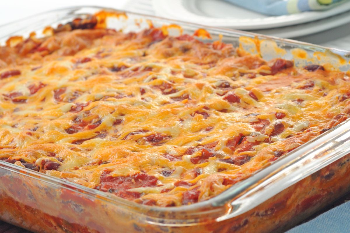 High protein Mexican beef taco casserole baked in a clear glass baking dish with layers of seasoned ground beef, tortillas, and tomato sauce. The top is evenly covered with melted shredded cheese, lightly browned and bubbly, with red sauce and beef visible along the edges in a kitchen setting.