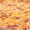 High protein Mexican beef taco casserole baked in a clear glass baking dish with visible layers of seasoned ground beef, tortillas, tomato sauce, and evenly melted shredded cheese. The top is golden and bubbly with red sauce peeking through, shown in a close-up food photography style.