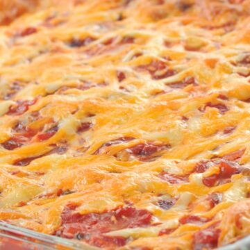 High protein Mexican beef taco casserole baked in a clear glass baking dish with visible layers of seasoned ground beef, tortillas, tomato sauce, and evenly melted shredded cheese. The top is golden and bubbly with red sauce peeking through, shown in a close-up food photography style.