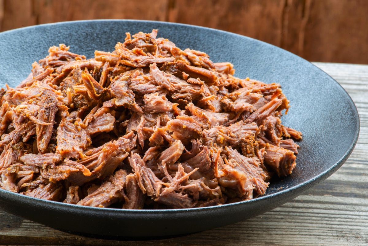 High protein orange and cumin pulled pork served in a dark shallow bowl on a rustic wooden table. Tender shredded pork coated in warm orange and cumin spices with a juicy, slow cooked texture. Savory pulled pork ready for bowls, wraps, or meal prep.